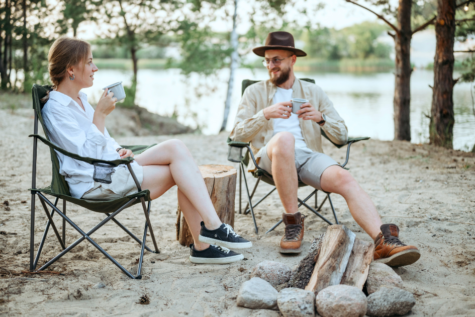 A couple sits relaxed in camp chairs a their site drinking a warm beverage. 