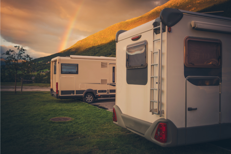 Two travel trailers parked side by side in a grassy spot with a sunset and rainbow in the background