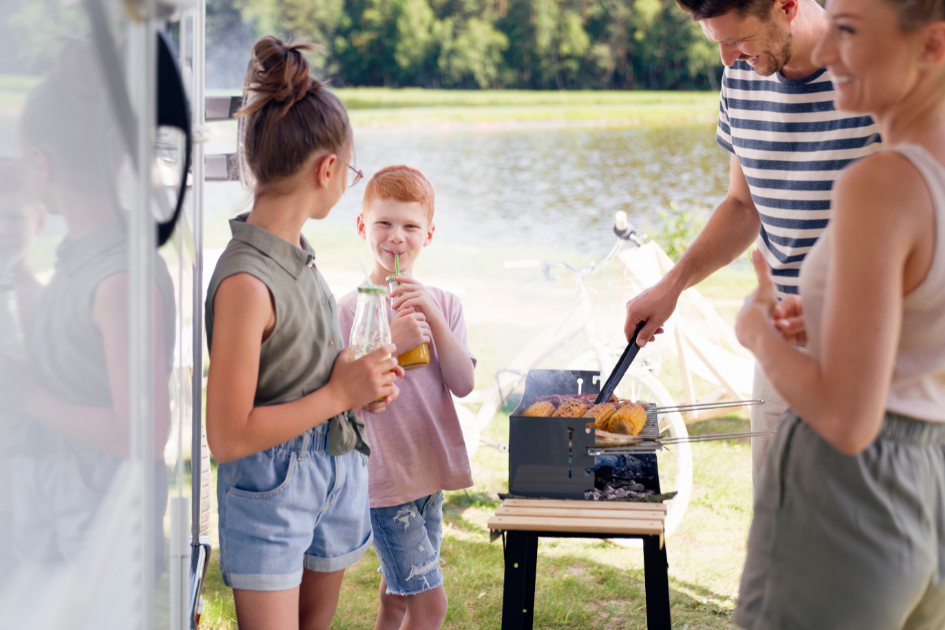 A father cooks lunch on a grill outside their RV, surrounded by his family