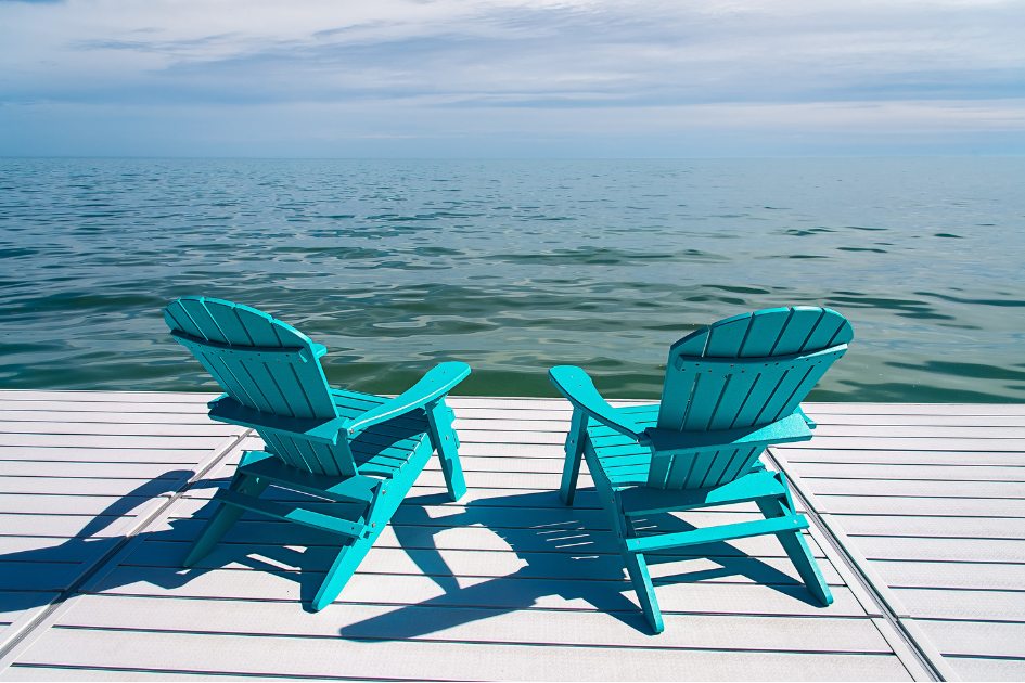 A pair of Adirondack chairs painted cyan blue sit o a dock overlooking a lake