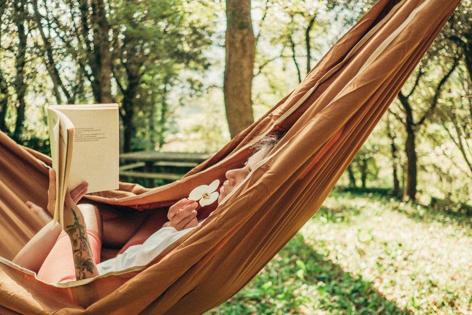 A person lays in a hammock in the shade of the trees, reading a book.