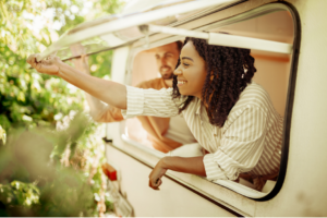 A woman gently leans out of her open RV window with a man smiling behind her.