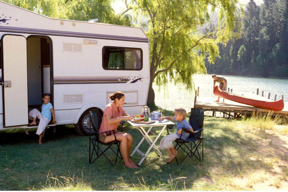 A mother and son sits at a picnic table while her daughter relaxes in the doorway of their RV and the dad gets a canoe ready at the dock nearby