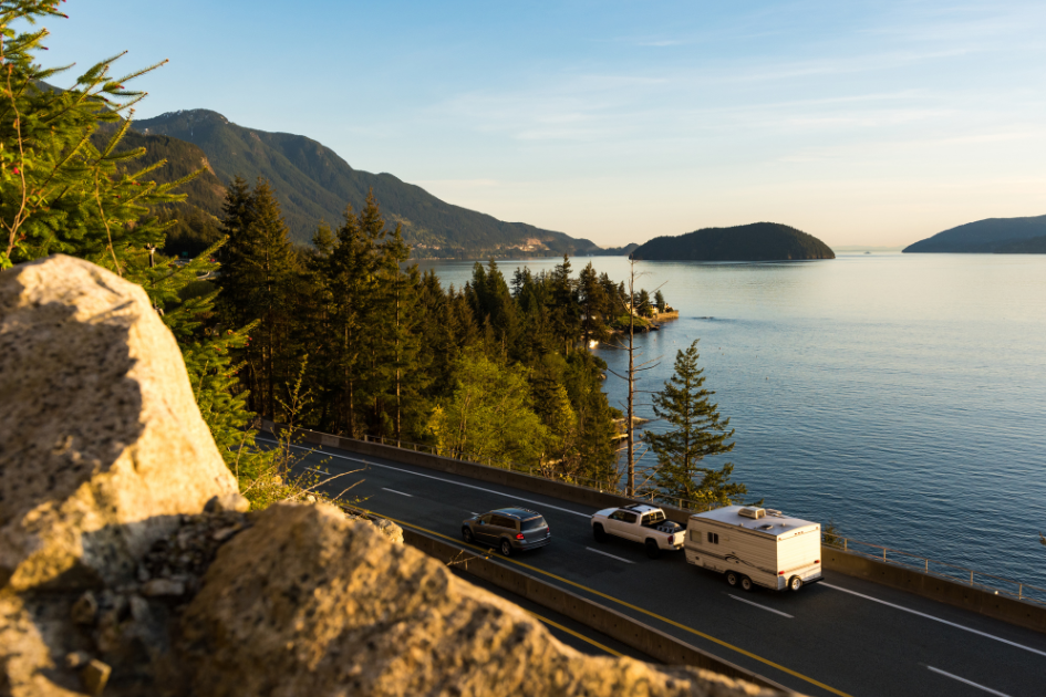 Truck towing a travel trailer behind it, driving down a scenic road next to a body of water with  mountains in the distance 