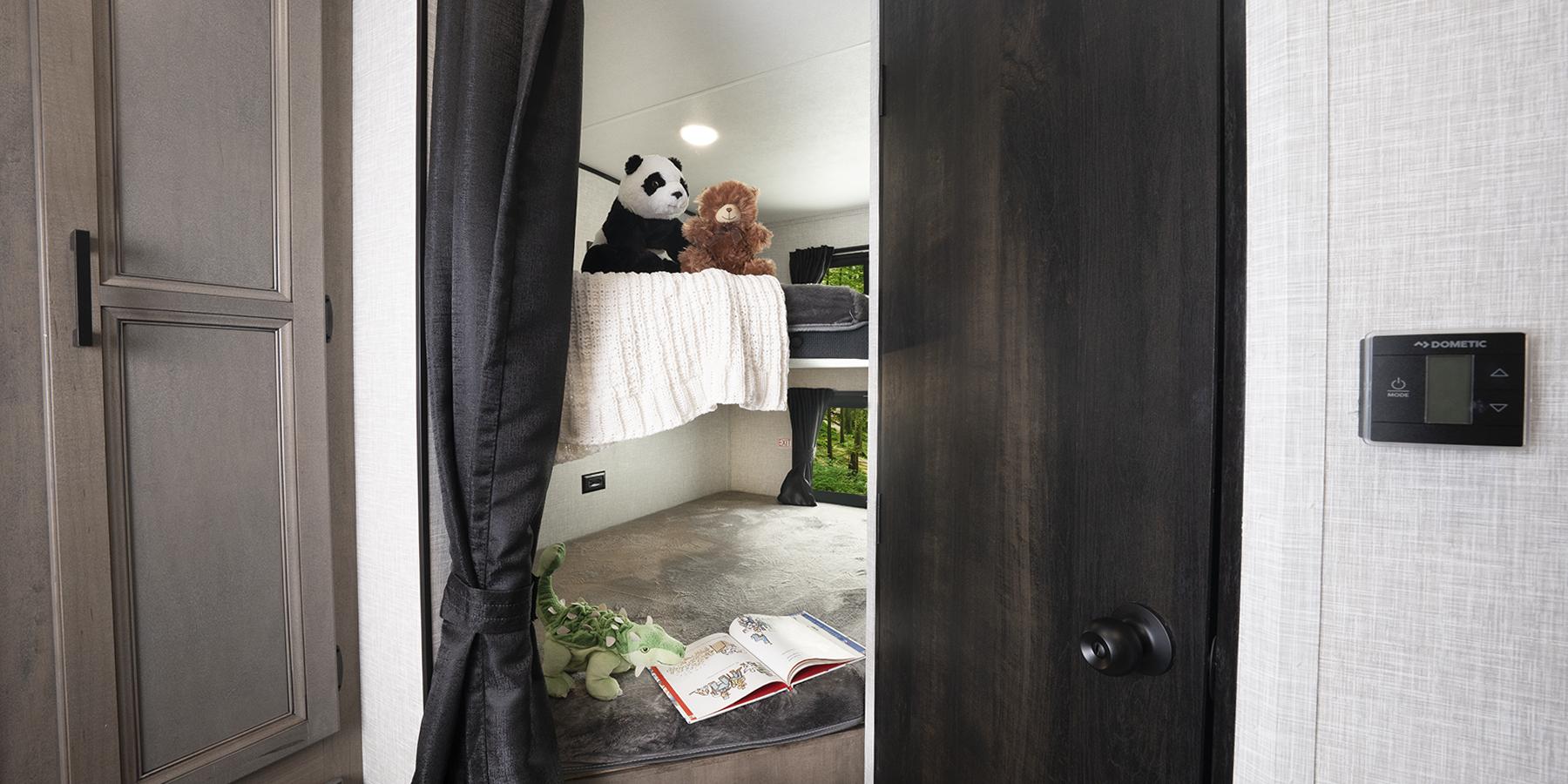 Through an open doorway with a drawn curtain tied off to one side, two double-sized bunks are visible, with childrens' books and stuffed animals neatly placed on top of the covers.